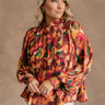 Front view of model wearing the Wrenley Multi Long Sleeve Top which features brown, orange, pink, green, purple, mustard, fuchsia and tan sheer fabric, pleated fabric, geomertic design, tan lining, high neckline with ruffle details, back key hole with but