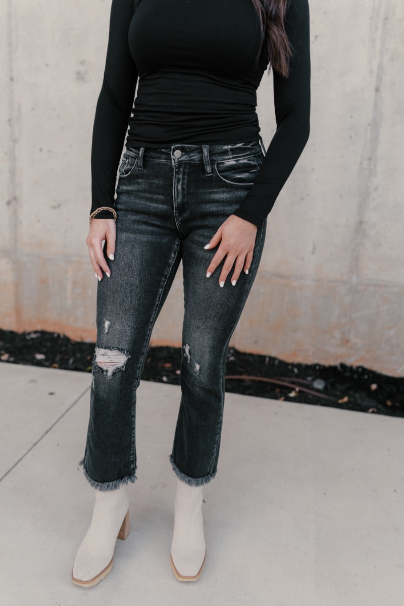 Front view of model wearing the Rooted Denim: Sydney Black Distressed Jeans that have distressed details, a front zipper belt loops, pockets and cropped flare legs with fray hem.