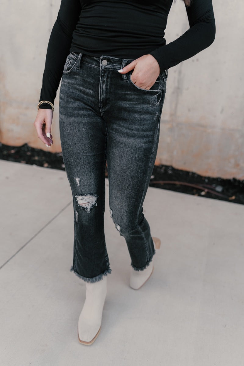 Front view of model wearing the Rooted Denim: Sydney Black Distressed Jeans that have distressed details, a front zipper belt loops, pockets and cropped flare legs with fray hem.