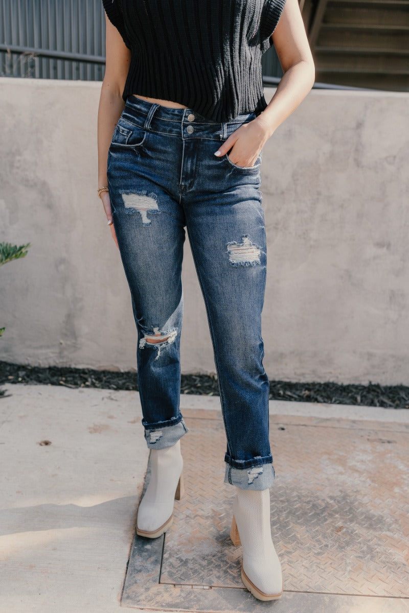 front view of model wearing the Rooted Denim: Kali Dark Wash Distressed Jeans that have dark wash denim, distressing, a front zipper and two-button closure, pockets, belt loops, and straight legs