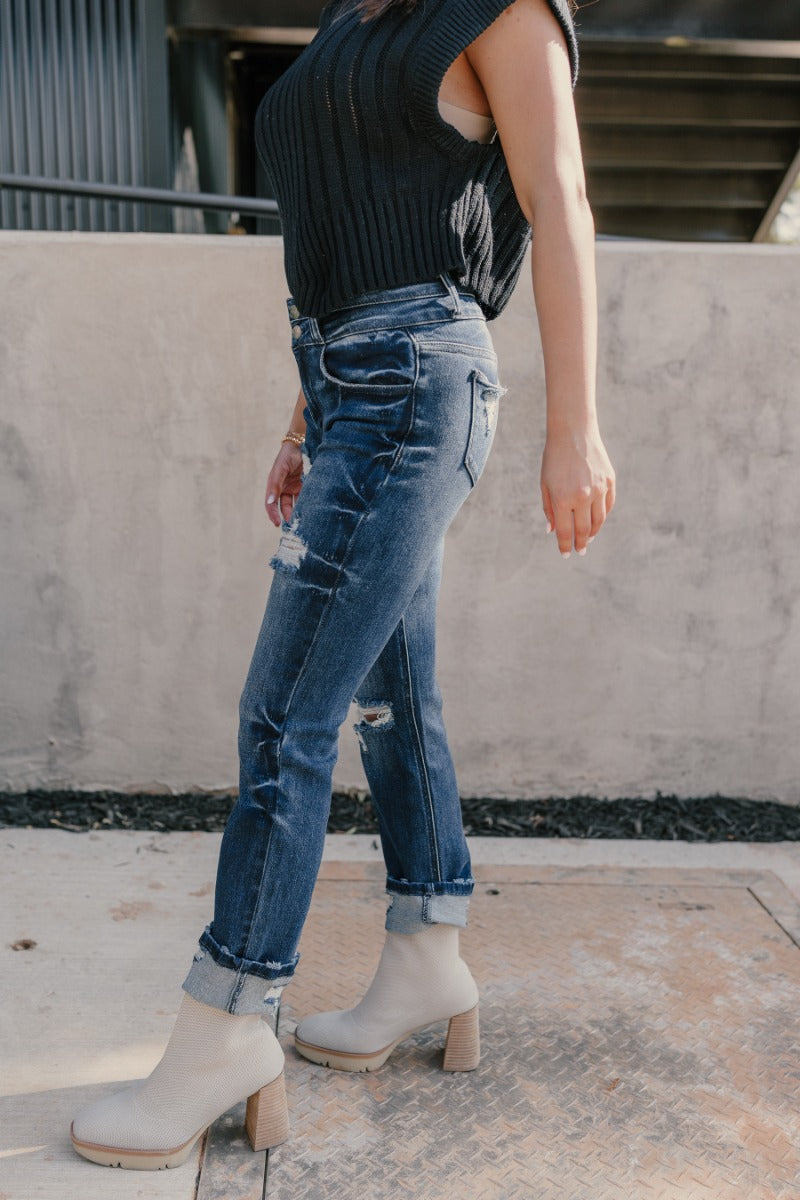 Side view of model wearing the Rooted Denim: Kali Dark Wash Distressed Jeans that have dark wash denim, distressing, a front zipper and two-button closure, pockets, belt loops, and straight legs