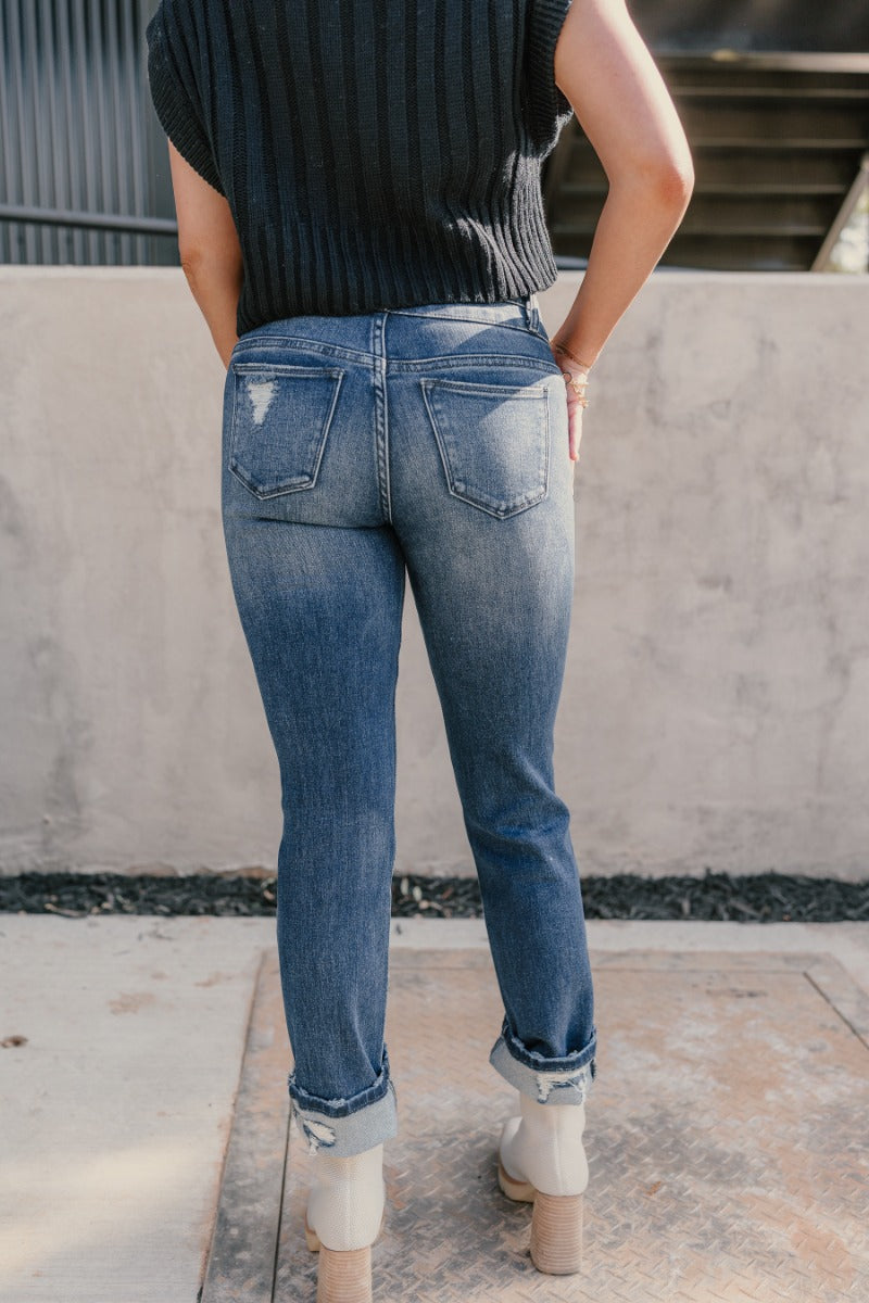 Back view of model wearing the Rooted Denim: Kali Dark Wash Distressed Jeans that have dark wash denim, distressing, a front zipper and two-button closure, pockets, belt loops, and straight legs