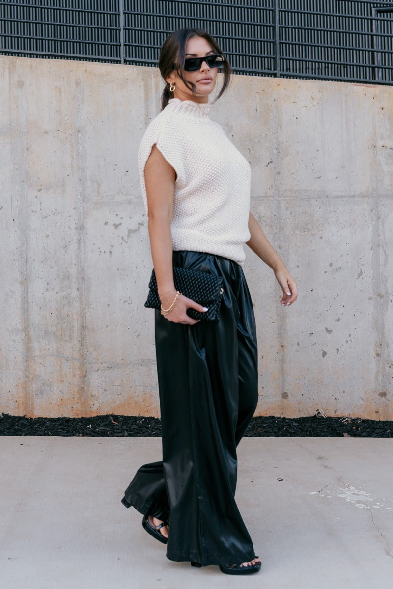 Full body side view of model wearing Malia Black Faux-Leather Wide Leg Pantsthat have feature black faux leather fabric, pockets, upper pleating, and wide legs.