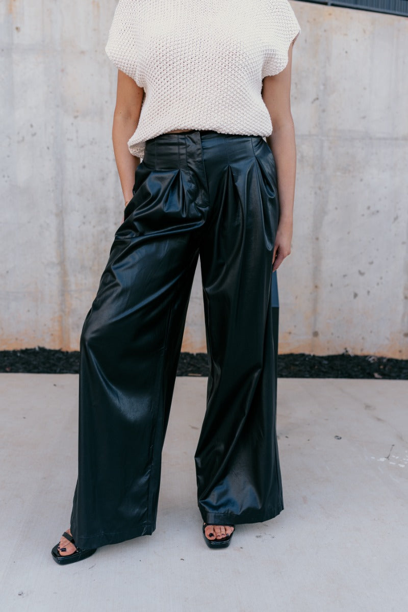 front view of model wearing Malia Black Faux-Leather Wide Leg Pantsthat have feature black faux leather fabric, pockets, upper pleating, and wide legs.