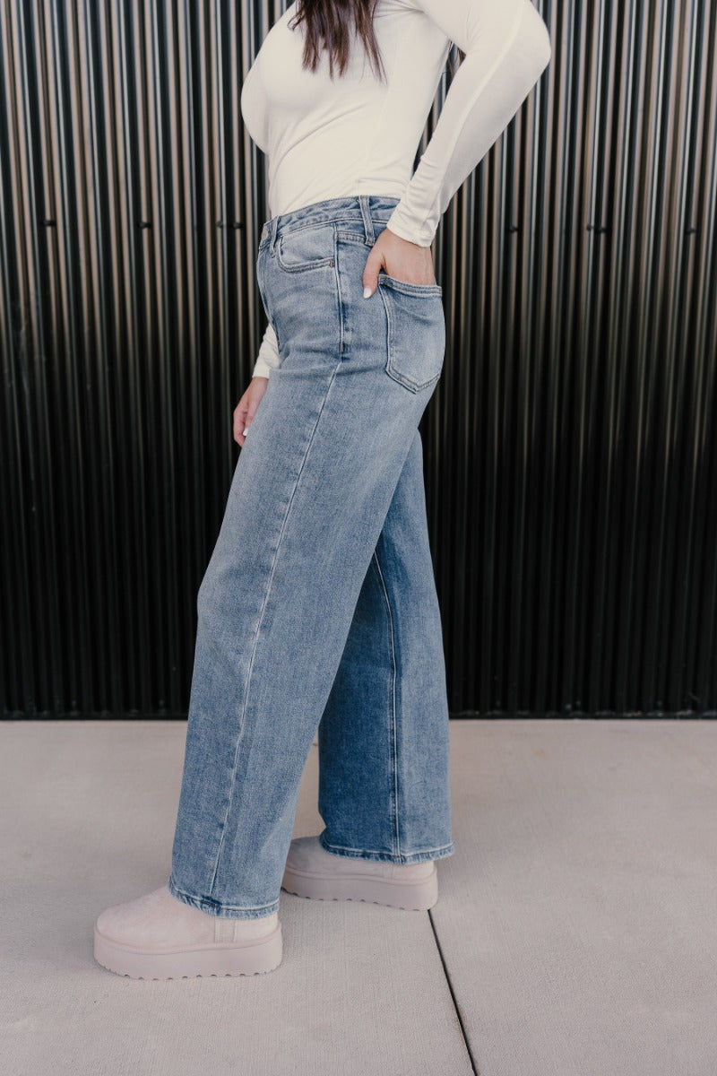 Side view of model wearing the Ceros: Daniela Wide Leg Jeans that have medium blue denim fabric, a front zipper, pockets, belt loops and wide legs.