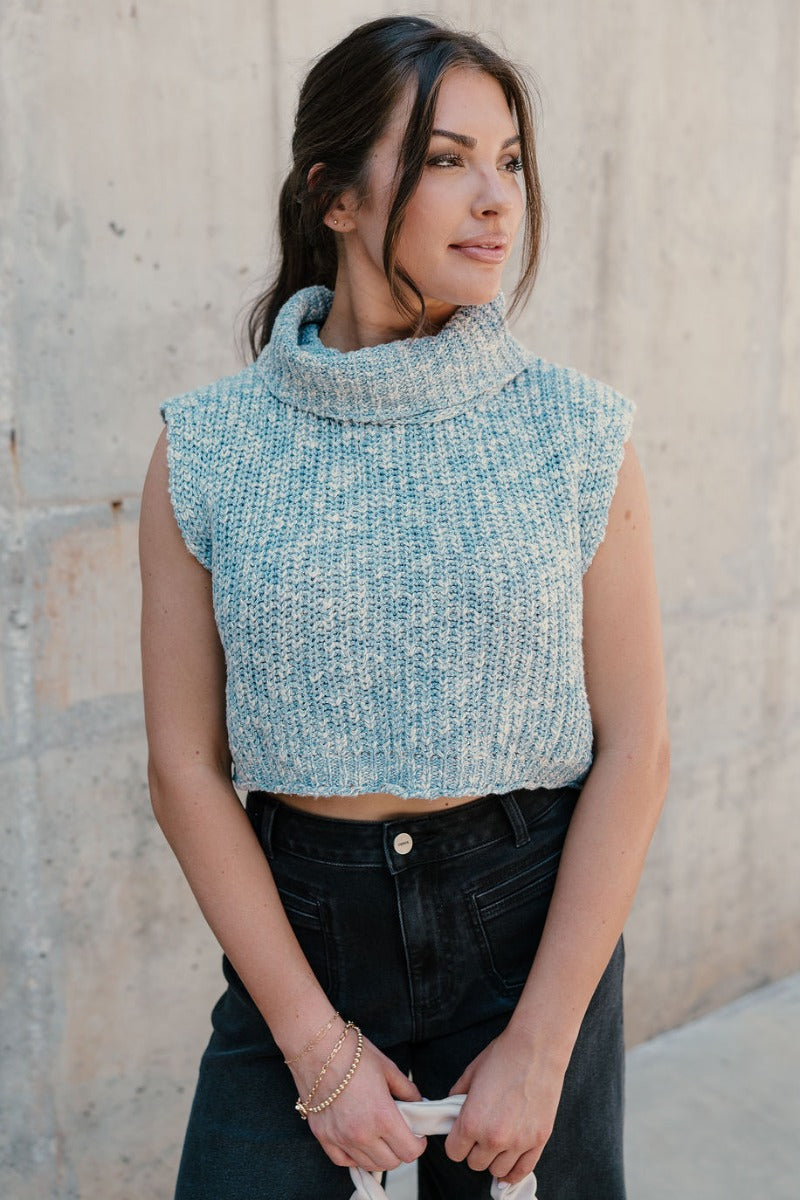 Front view of model wearing the Jordyn Blue & White Sleeveless Turtleneck Sweater which features blue and white cable knit fabric, a cropped waist, a turtleneck neckline, and a sleeveless design.