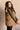 Side view of model wearing the Esme Chartreuse Ruffle Puffer Vest, that has olive puffer fabric, button up closures, two front pockets, high collar neck, short sleeves with ruffles. Worn unbuttoned over cream dress.