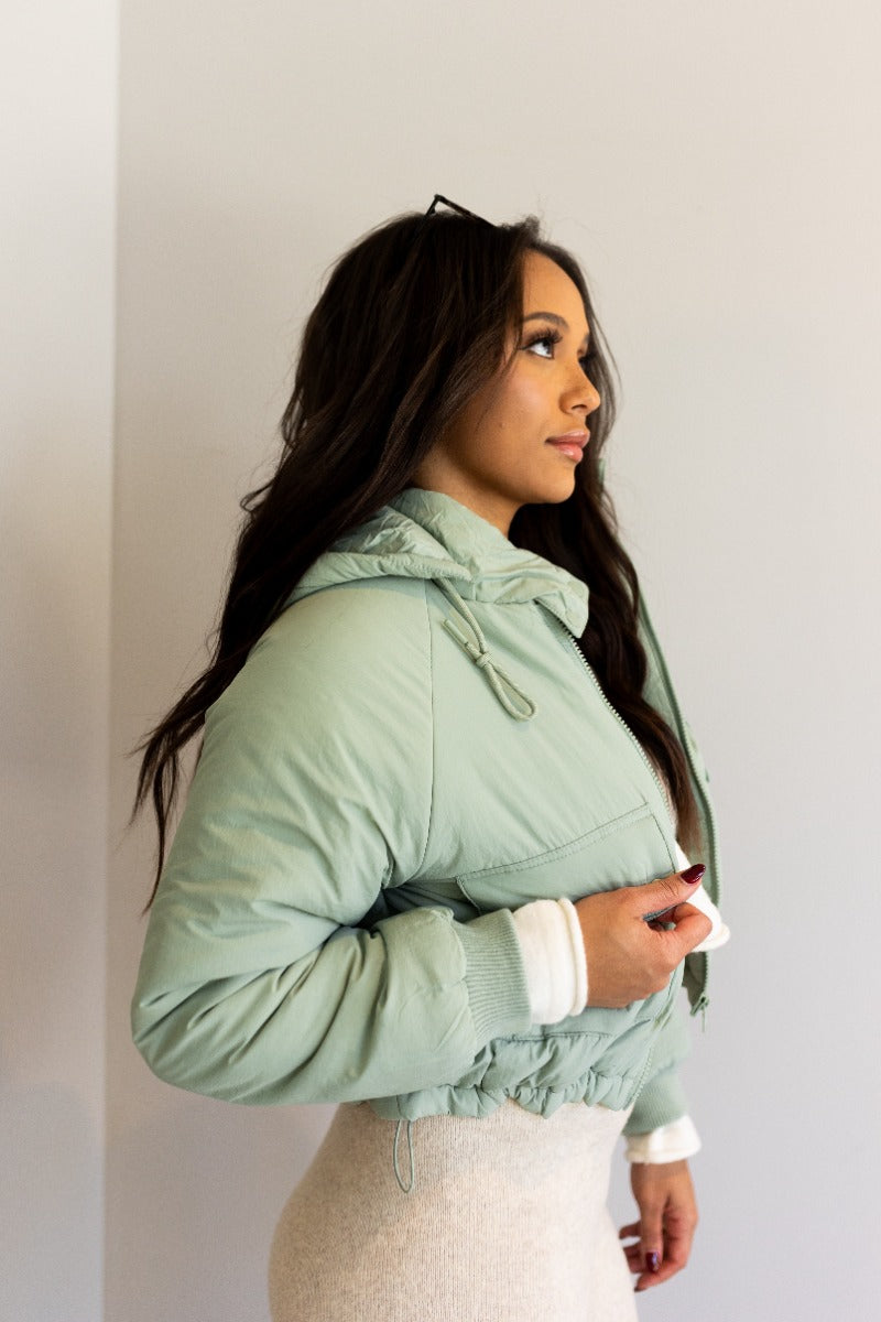 Side view of model wearing the Mckinley Mint Cropped Puffer Jacket that has mint green puffer fabric, a cropped waist, a front zipper with snaps, a hood, and long sleeves.