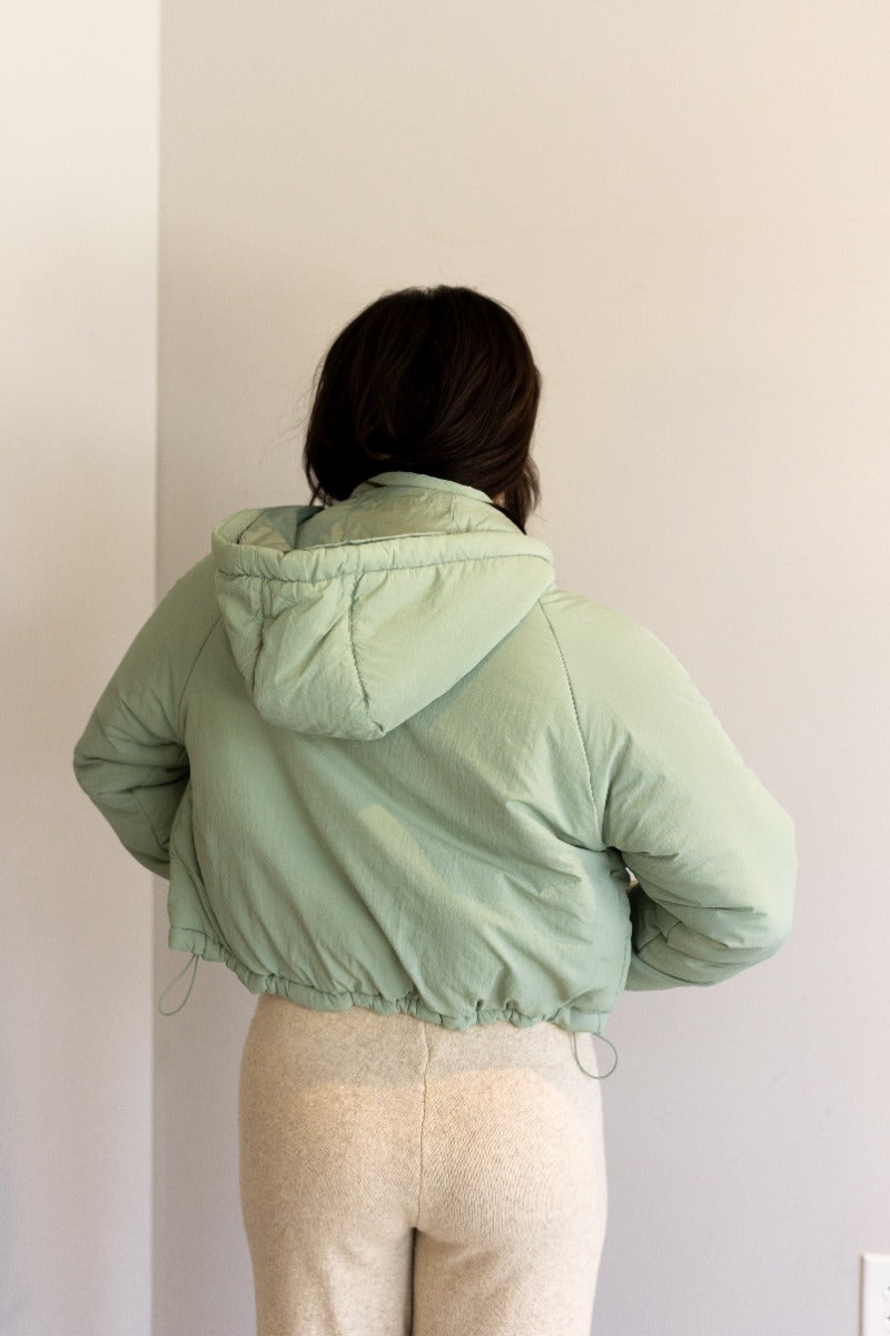 Back view of model wearing the Mckinley Mint Cropped Puffer Jacket that has mint green puffer fabric, a cropped waist, a front zipper with snaps, a hood, and long sleeves.