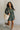 Full body front view of model wearing the Ezra Grey Green Long Sleeve Romper that has grey green knit fabric, pockets, an elastic waist, a front zipper, a hood, a skirt with fitted shorts , and long sleeves.
