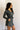 Side view of model wearing the Ezra Grey Green Long Sleeve Romper that has grey green knit fabric, pockets, an elastic waist, a front zipper, a hood, a skirt with fitted shorts , and long sleeves.
