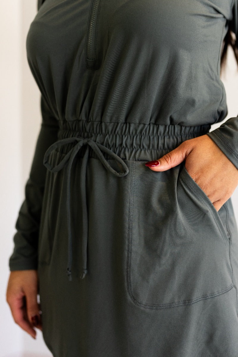 Close front view of model wearing the Ezra Grey Green Long Sleeve Romper that has grey green knit fabric, pockets, an elastic waist, a front zipper, a hood, a skirt with fitted shorts , and long sleeves.