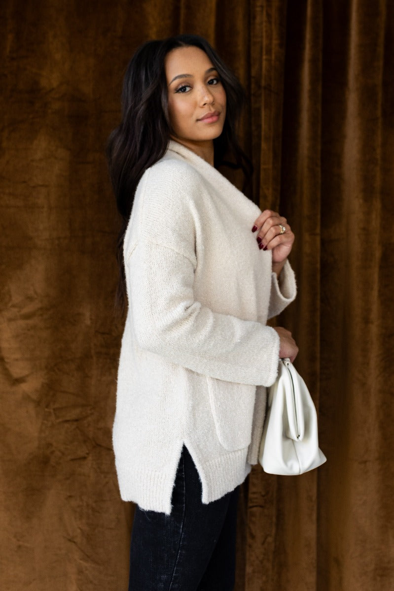 Side view of model wearing the Elena Beige Open Front Cardigan that has beige knit fabric, two front pockets, ribbed trim , an open front, and long sleeves.