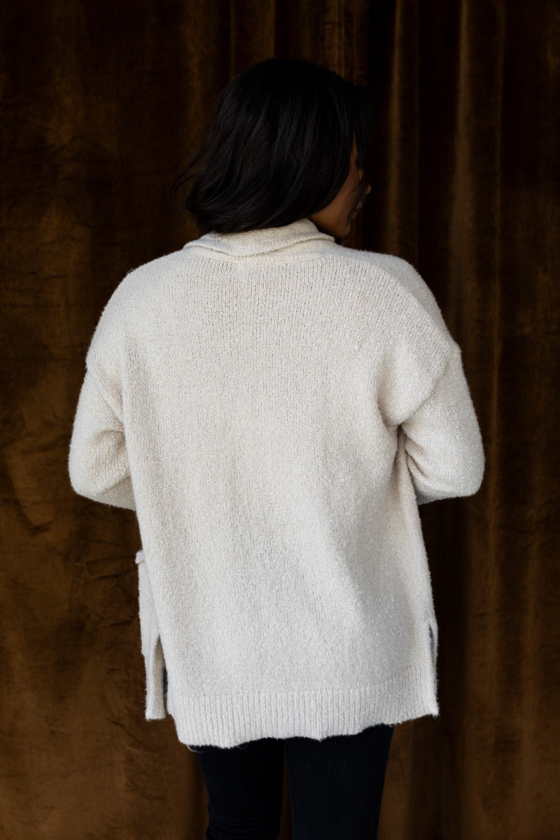 Back view of model wearing the Elena Beige Open Front Cardigan that has beige knit fabric, two front pockets, ribbed trim , an open front, and long sleeves.