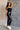 Full body side view of model wearing the Vera Black Sleeveless Bodysuit which features black knit fabric, v-neckline, adjustable spaghetti straps, sleeveless and thong bottom with button snap closure.