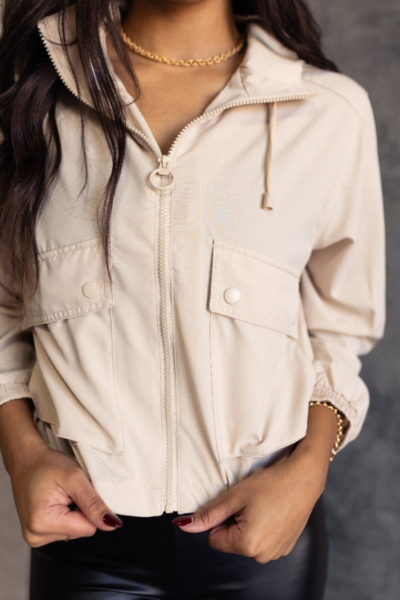 Close up view of model wearing the Leia Stone Lightweight Long Sleeve Jacket which features beige lightweight fabric, drawstring hem, monochromatic front zip up, two front buttoned pockets, hoode attached and long sleeves with elastic wrists.