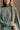 Detail view of model wearing The Jennifer Sage Mock Neck Sweatshirt features sage green knit fabric, two front slit pockets, ribbed hem, high neckline and long sleeves with cuffs.