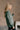 Side view of model wearing The Jennifer Sage Mock Neck Sweatshirt features sage green knit fabric, two front slit pockets, ribbed hem, high neckline and long sleeves with cuffs.
