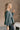 Side view of model wearing The Bella Pine Green Long Sleeve Top features pine green gauze fabric, peplum body, round neckline, long puff sleeves with elastic cuffs and open back with tie closure.