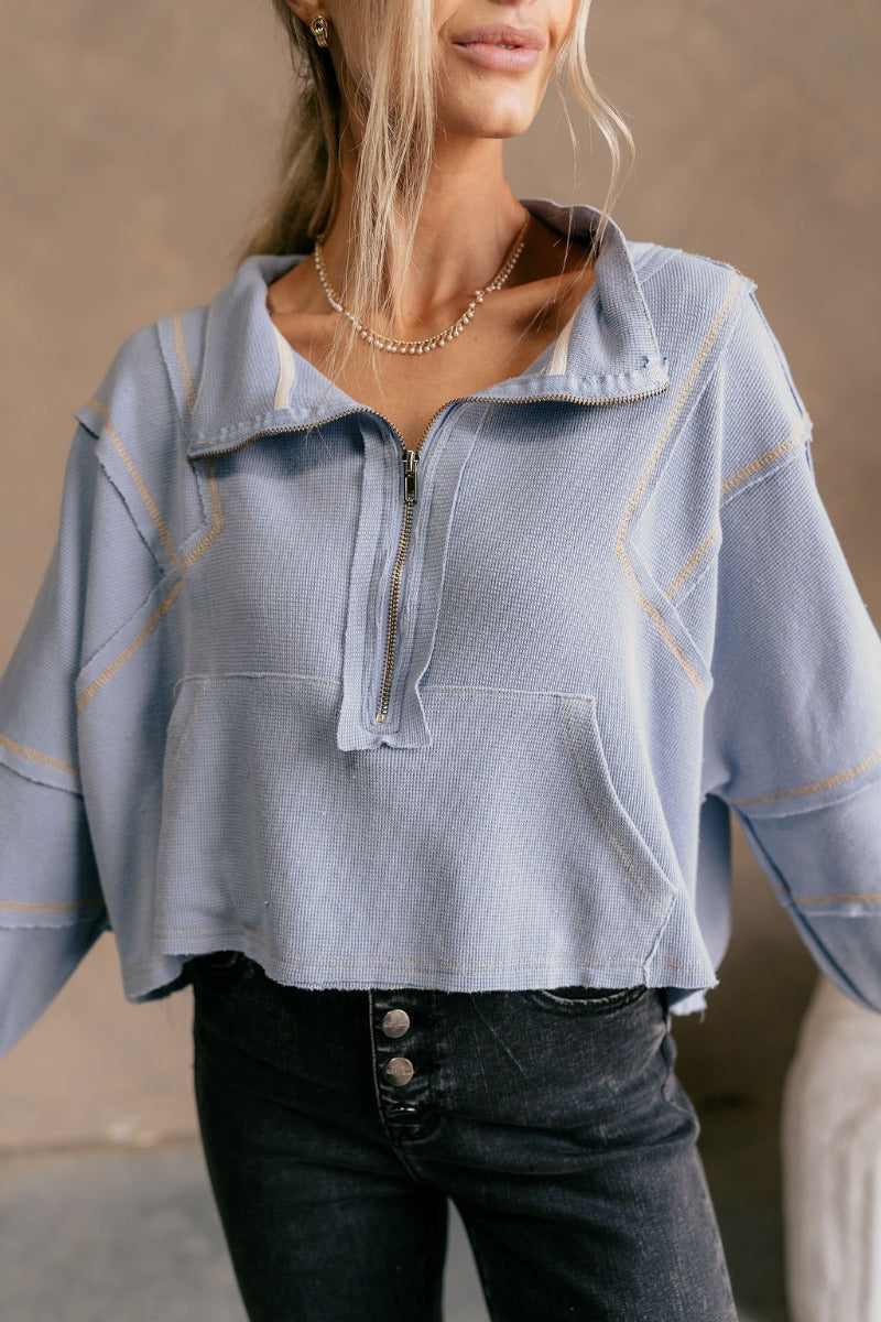Front view of model wearing The Anna Periwinkle Thermal Long Sleeve Top features periwinkle blue knit fabric, light mustard threading, cream stripe design on the sleeves, cropped waist, one front pocket, quarter zip up, high neckline, distressed details a