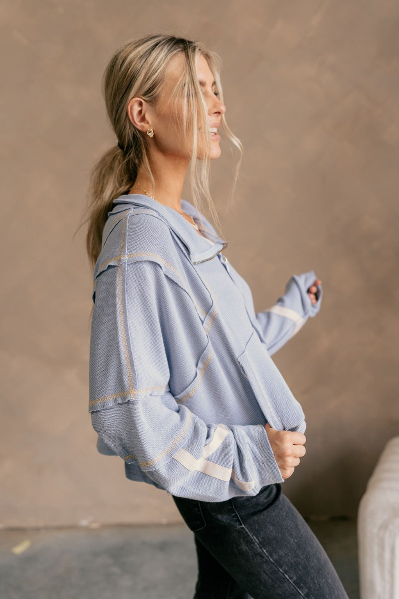Side view of model wearing The Anna Periwinkle Thermal Long Sleeve Top features periwinkle blue knit fabric, light mustard threading, cream stripe design on the sleeves, cropped waist, one front pocket, quarter zip up, high neckline, distressed details an