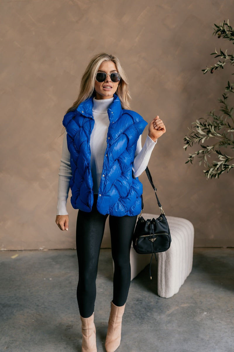 Front view of model wearing The Laurel Royal Blue Button-Up Puffer Vest features cobalt blue nylon puffer fabric, intertwined design, monochrome button up closure, high neckline and sleeveless.