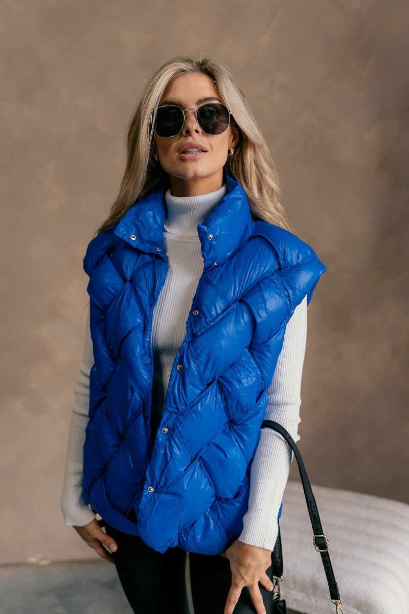 Front view of model wearing The Laurel Royal Blue Button-Up Puffer Vest features cobalt blue nylon puffer fabric, intertwined design, monochrome button up closure, high neckline and sleeveless.