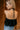 Back view of model wearing the Priscilla Black Rose Detail Halter Top that has black lightweight fabric, black lining, a cropped waist, a halter strap tie neckline, and a dramatic black rosette detail.