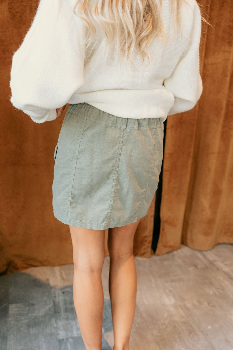 Back view of model wearing The Sage Corduroy Cargo Skirt features sage textured fabric, two front cargo pockets, mini length, belt loops and front zipper with green tortoise button closure.