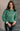 Front view of model wearing the Merry Green Ribbed Long Sleeve Sweater which features green and light green ribbed fabric, thick hem, a round neckline, a graphic that says "MERRY" in white outline, dropped shoulders, and long sleeves with ribbed cuffs.