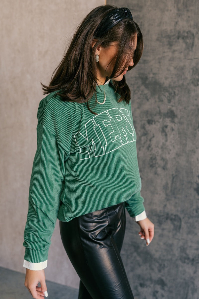 Side view of model wearing the Merry Green Ribbed Long Sleeve Sweater which features green and light green ribbed fabric, thick hem, a round neckline, a graphic that says "MERRY" in white outline, dropped shoulders, and long sleeves with ribbed cuffs.