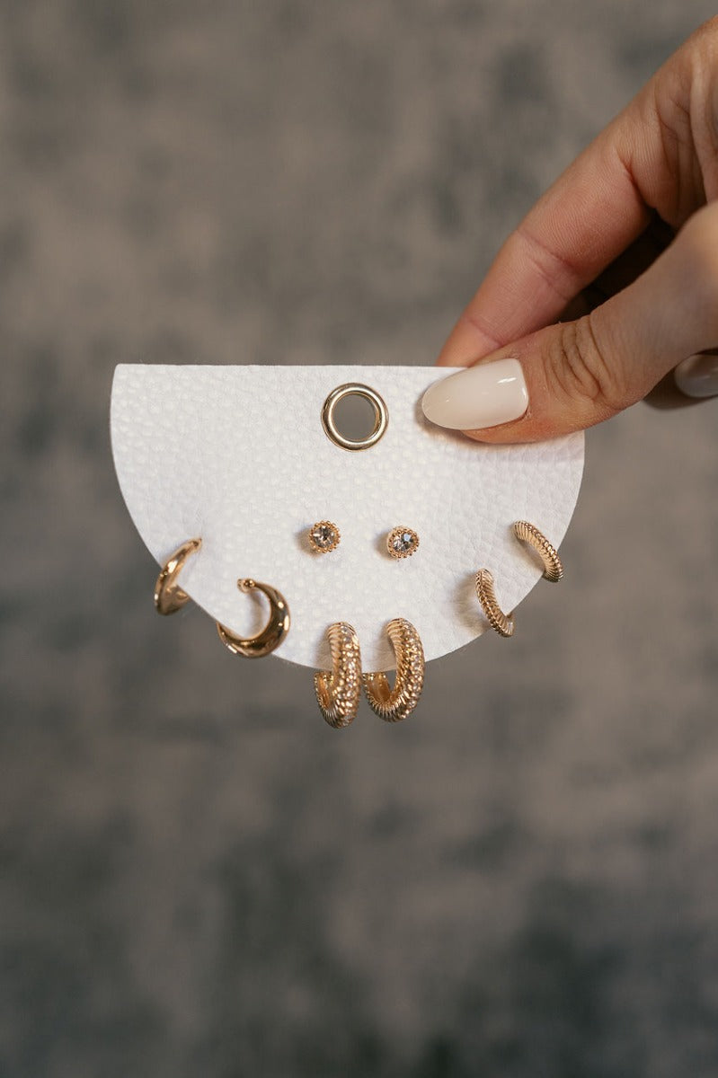 Close up view of model holding the Gold Hoops & Stud Earring Set which features four different pairs of earrings, one pair of small gold hoops, one pair of medium gold hoops with clear stones, one pair of small textured gold hoops and one pair of rhinesto