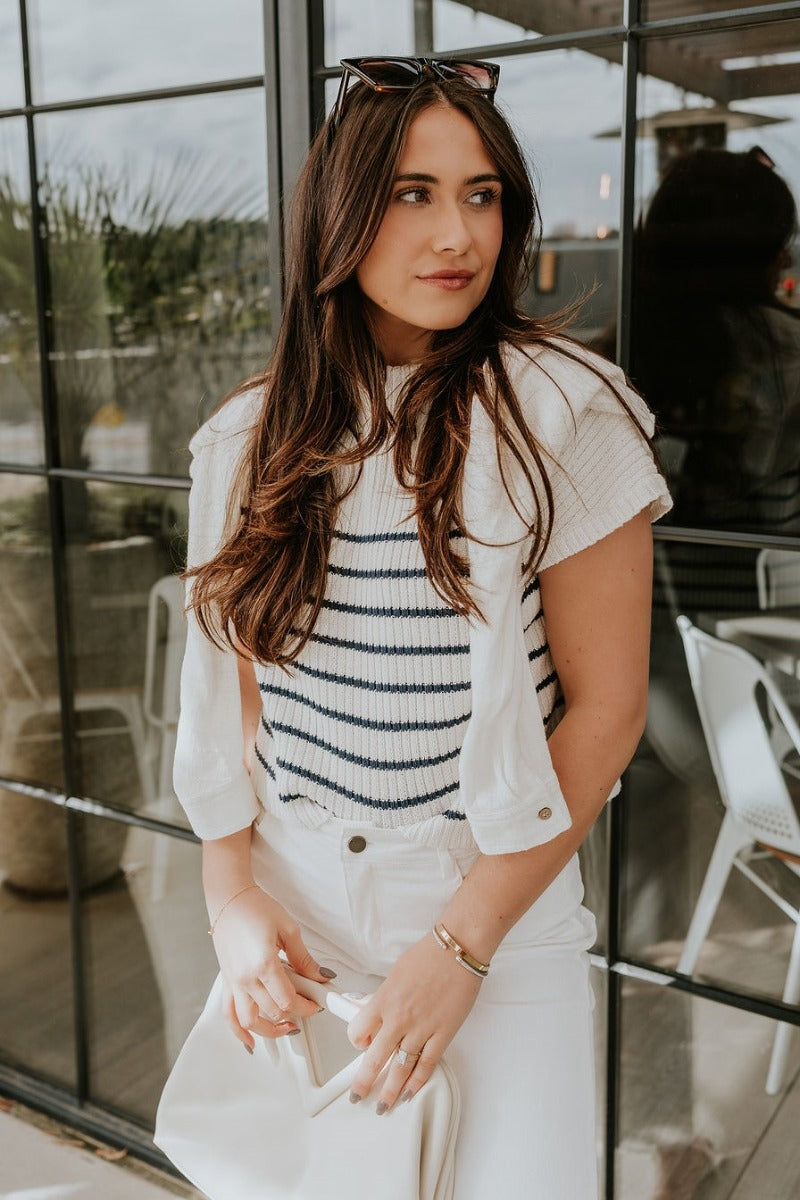 Front view of model wearing the Katelyn White & Navy Striped Sleeveless Sweater that has white and navy knit fabric, a striped pattern, a mock neckline, and a sleeveless design. Paired with white jacket.