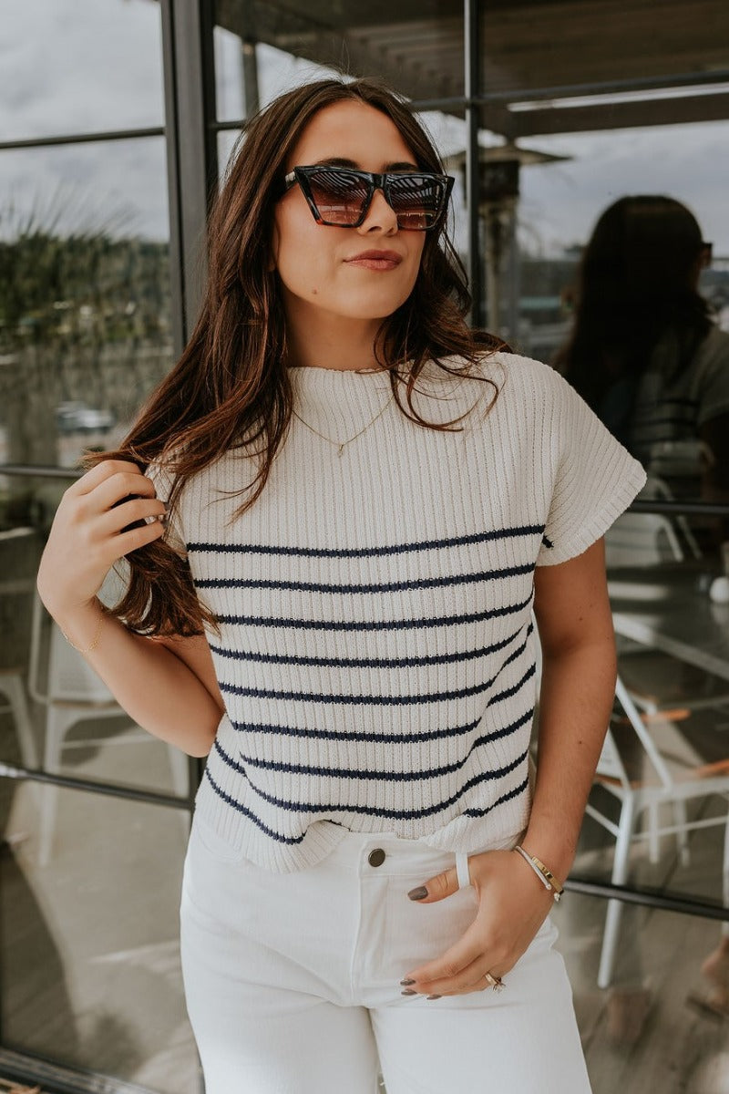 Front view of model wearing the Katelyn White & Navy Striped Sleeveless Sweater that has white and navy knit fabric, a striped pattern, a mock neckline, and a sleeveless design.