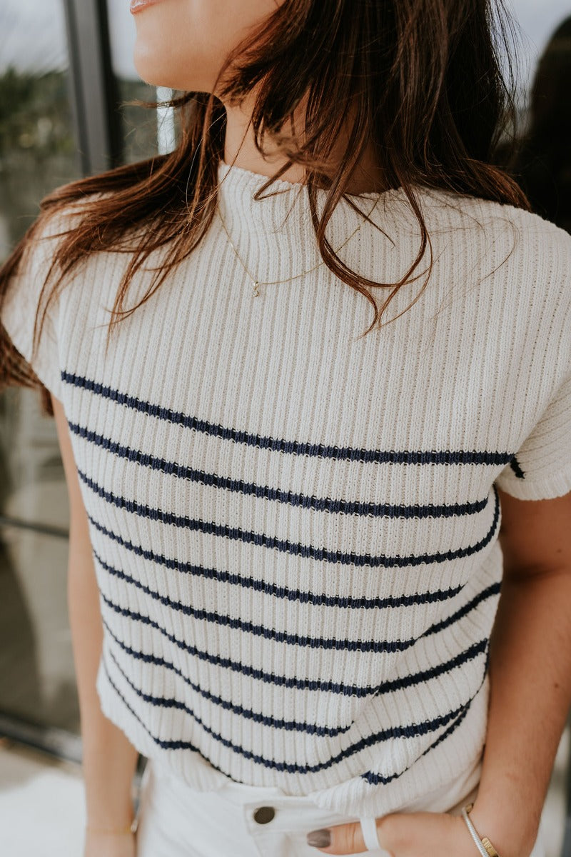 Close front view of model wearing the Katelyn White & Navy Striped Sleeveless Sweater that has white and navy knit fabric, a striped pattern, a mock neckline, and a sleeveless design.