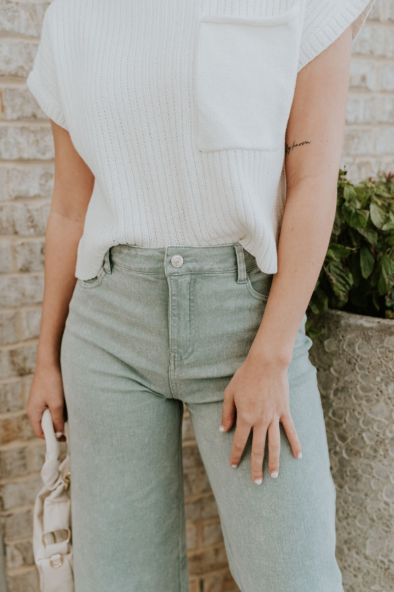Upper front view of model wearing the Livia Sage Wide Leg Pants that has sage green washed denim-like fabric, a front zipper with a button closure, belt loops, front pockets, back pockets and wide pant legs.