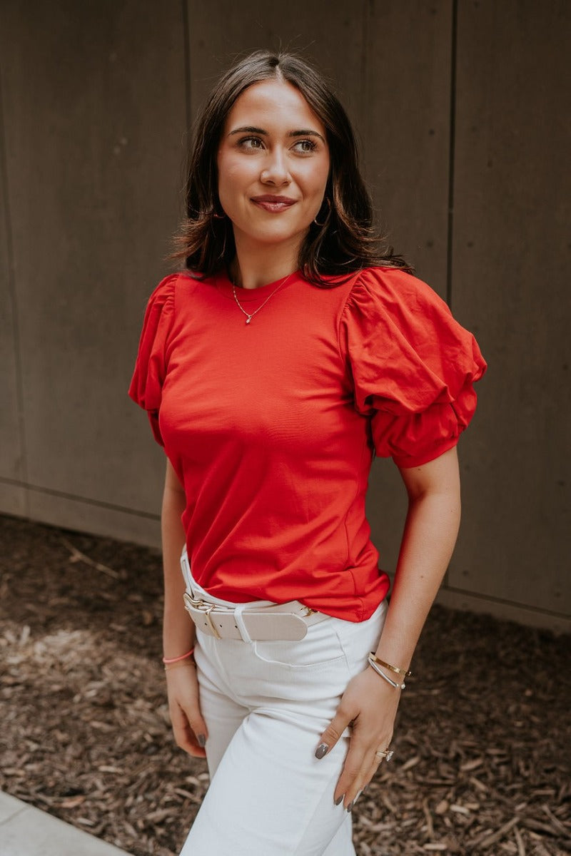 Frontal side view of model wearing the Maddie Red Puff Sleeve Top that features red cotton fabric, a round neckline, and short bubble puff sleeves.