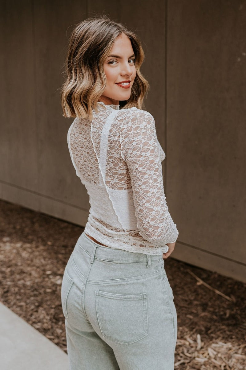 Side view of model wearing the Abby White Lace Long Sleeve Top that has white sheer fabric, monochrome lace floral pattern, lettuce hem, a high neckline, and long sleeves.