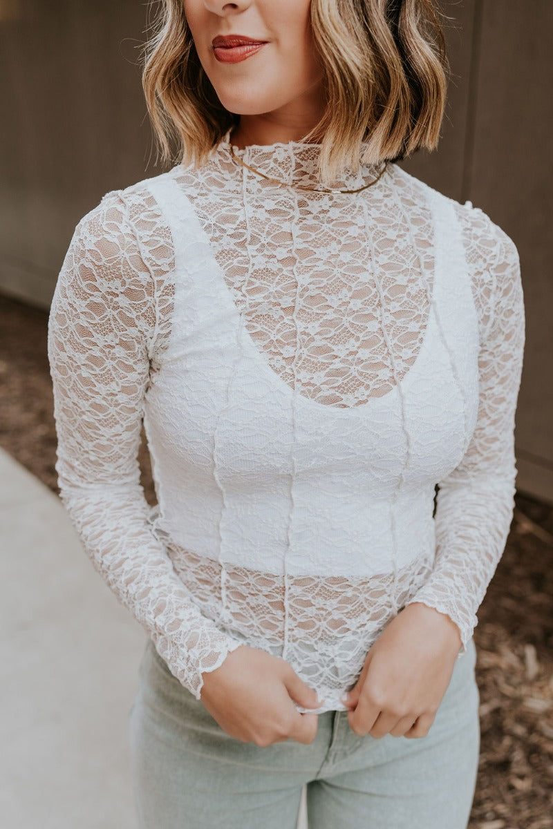 Close front view of model wearing the Abby White Lace Long Sleeve Top that has white sheer fabric, monochrome lace floral pattern, lettuce hem, a high neckline, and long sleeves.