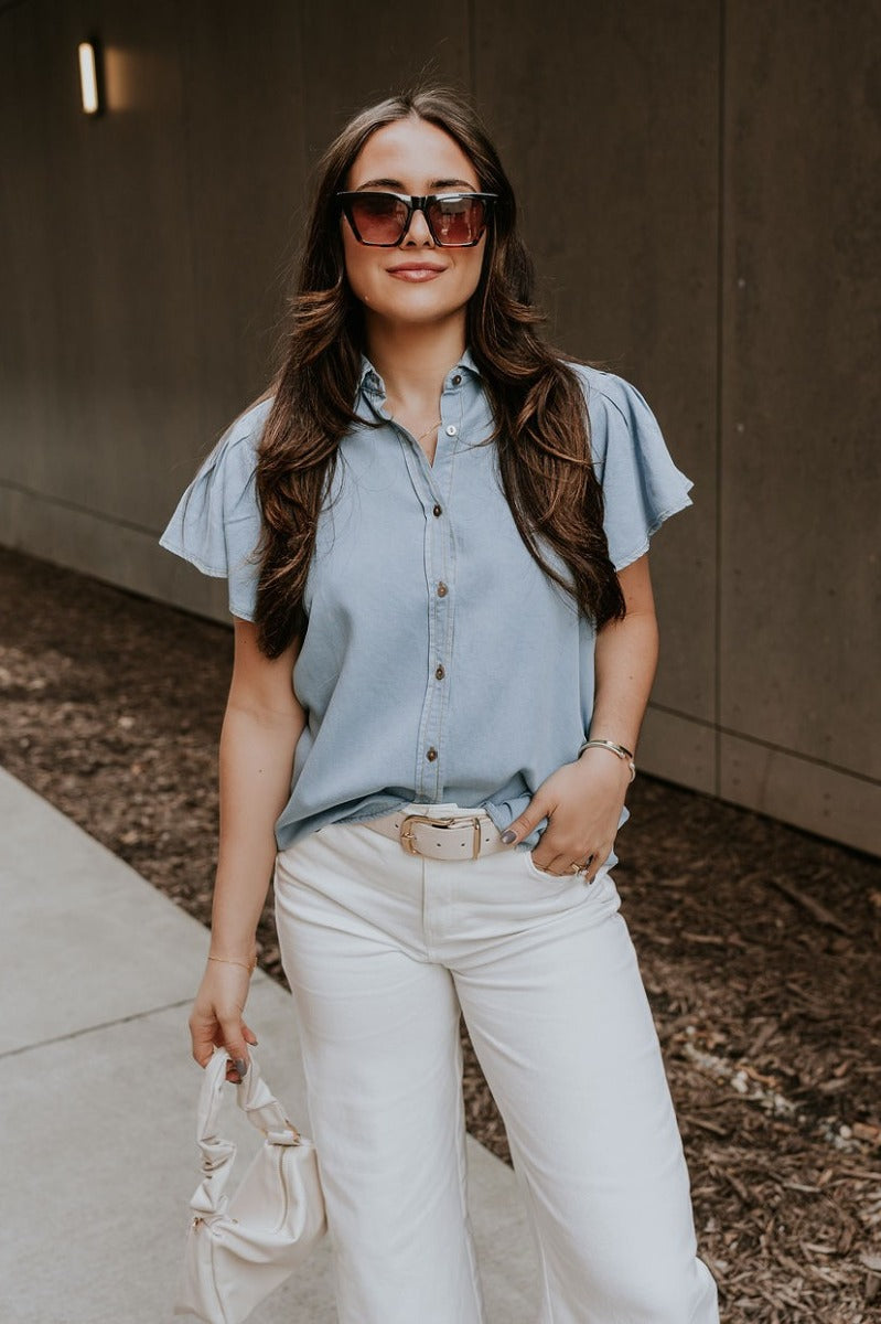 Front view of model wearing the Cynthia Blue Short Sleeve Button Up Top that has light blue tencel fabric, light brown stitching , grey tortoise buttons, a collared neckline, and short flare sleeves.