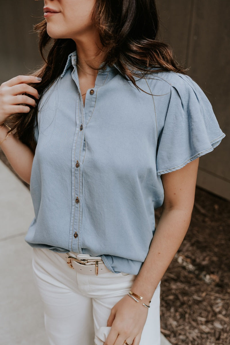Close front view of model wearing the Cynthia Blue Short Sleeve Button Up Top that has light blue tencel fabric, light brown stitching , grey tortoise buttons, a collared neckline, and short flare sleeves.