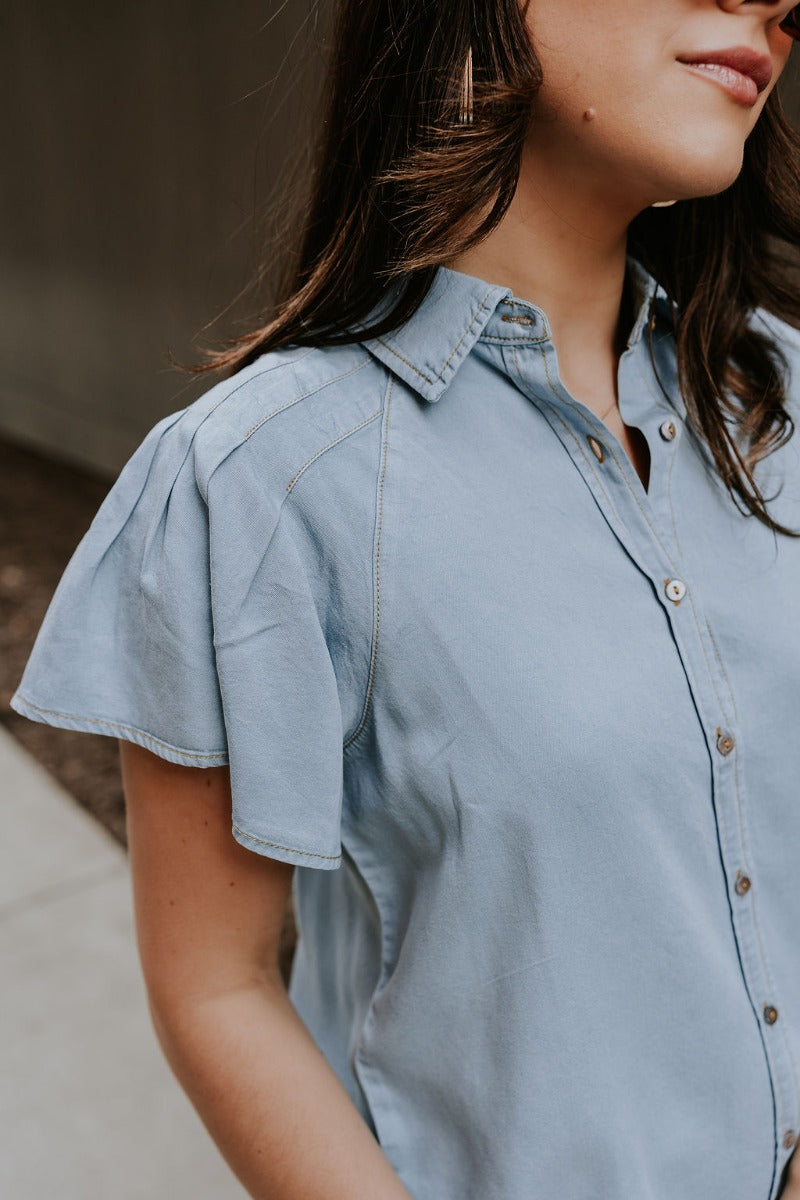 Upper front view of model wearing the Cynthia Blue Short Sleeve Button Up Top that has light blue tencel fabric, light brown stitching , grey tortoise buttons, a collared neckline, and short flare sleeves.