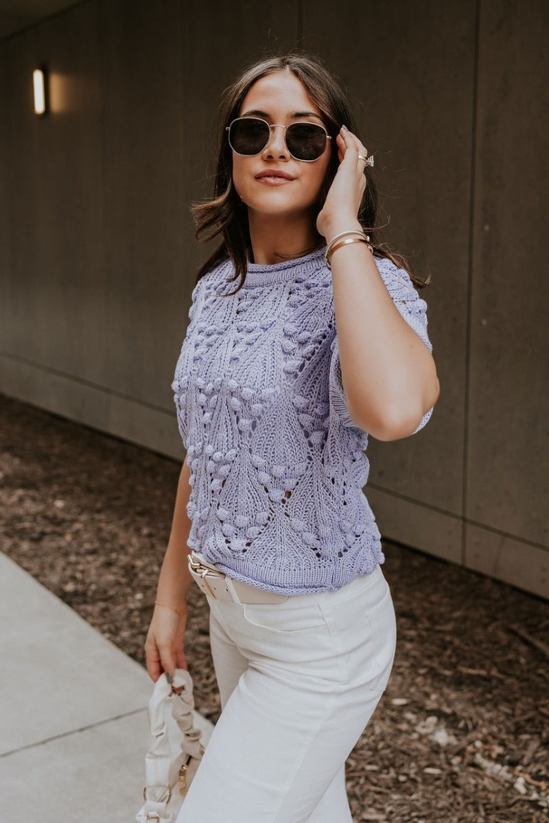Side view of model wearing the Megan Periwinkle Crochet Short Sleeve Sweater that has lavender-blue crochet knit fabric, monochrome 3d polka dot details, ribbed hem, a round neck, and short sleeves.