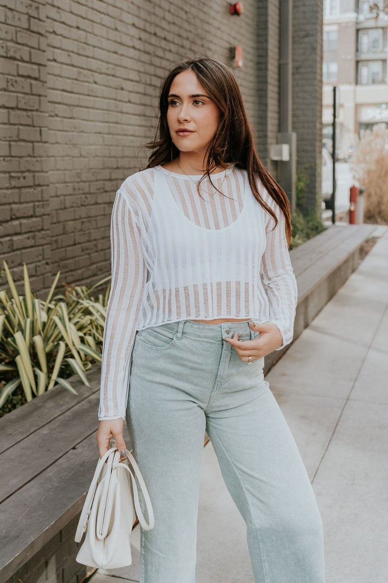 Front view of model wearing the Dalia White Open Knit Cropped Long Sleeve Top that has white open knit fabric, a monochrome stripe pattern, a round neck, a cropped waist, and long sleeves.