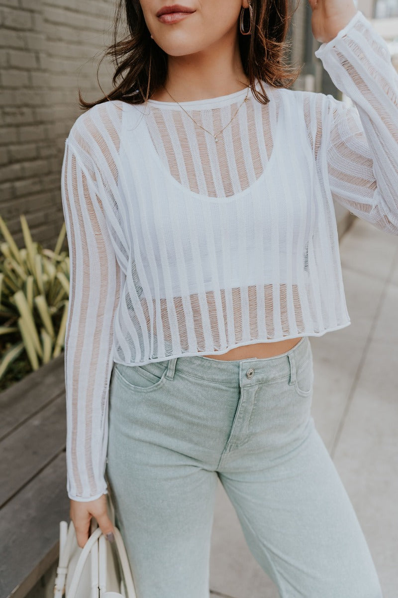 Front view of model wearing the Dalia White Open Knit Cropped Long Sleeve Top that has white open knit fabric, a monochrome stripe pattern, a round neck, a cropped waist, and long sleeves.