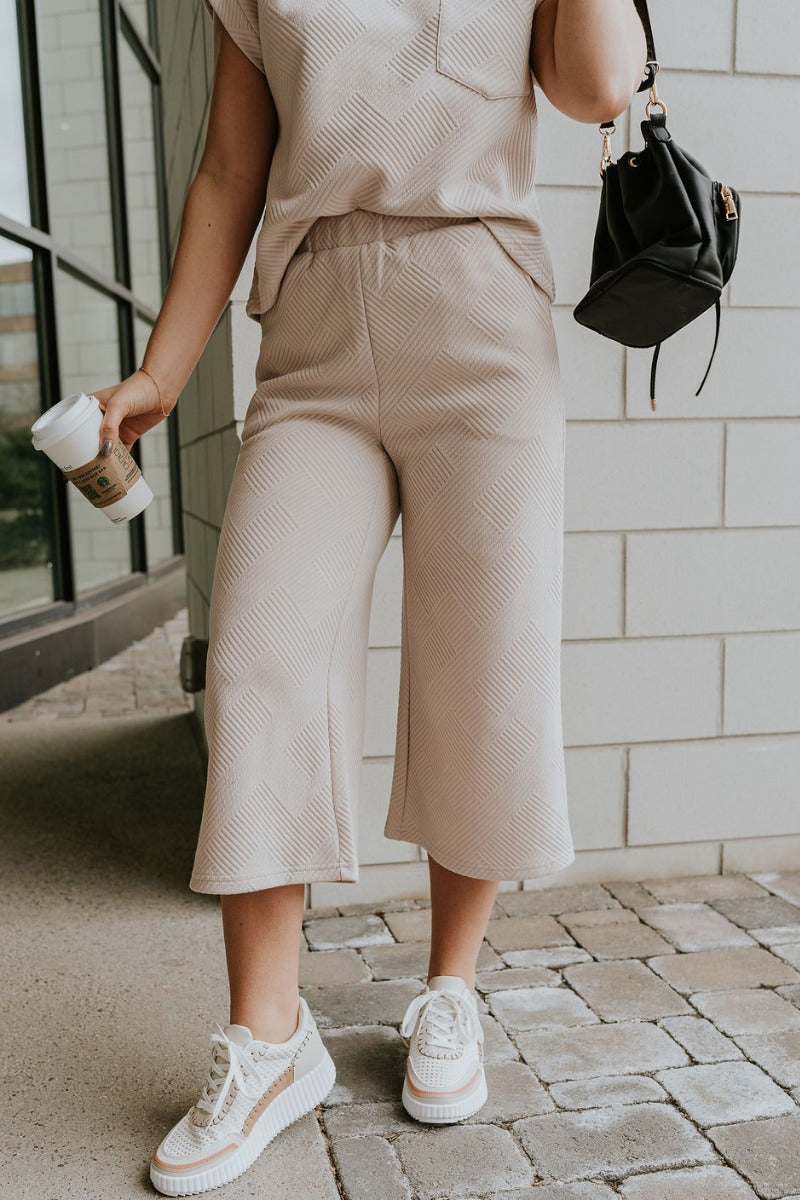 Front view of model wearing the Ella Light Taupe Cropped Pants which features light taupe knit fabric, monochrome block stripe design, elastic waistband, two front slit pockers and cropped wide pant legs.