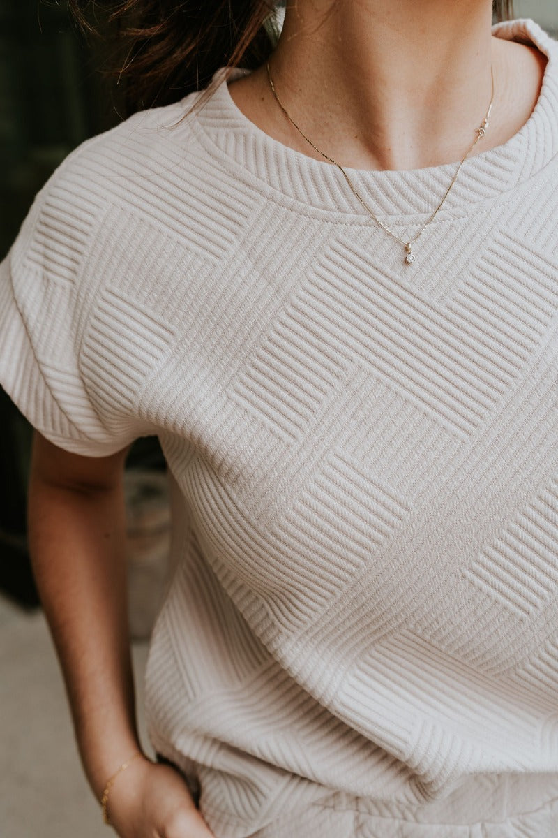 Close up view of model wearing the Ella Light Taupe Short Sleeve Top which features light taupe knit fabric, monochrome block stripe design, front left chest pocket, round neckline and short sleeves.