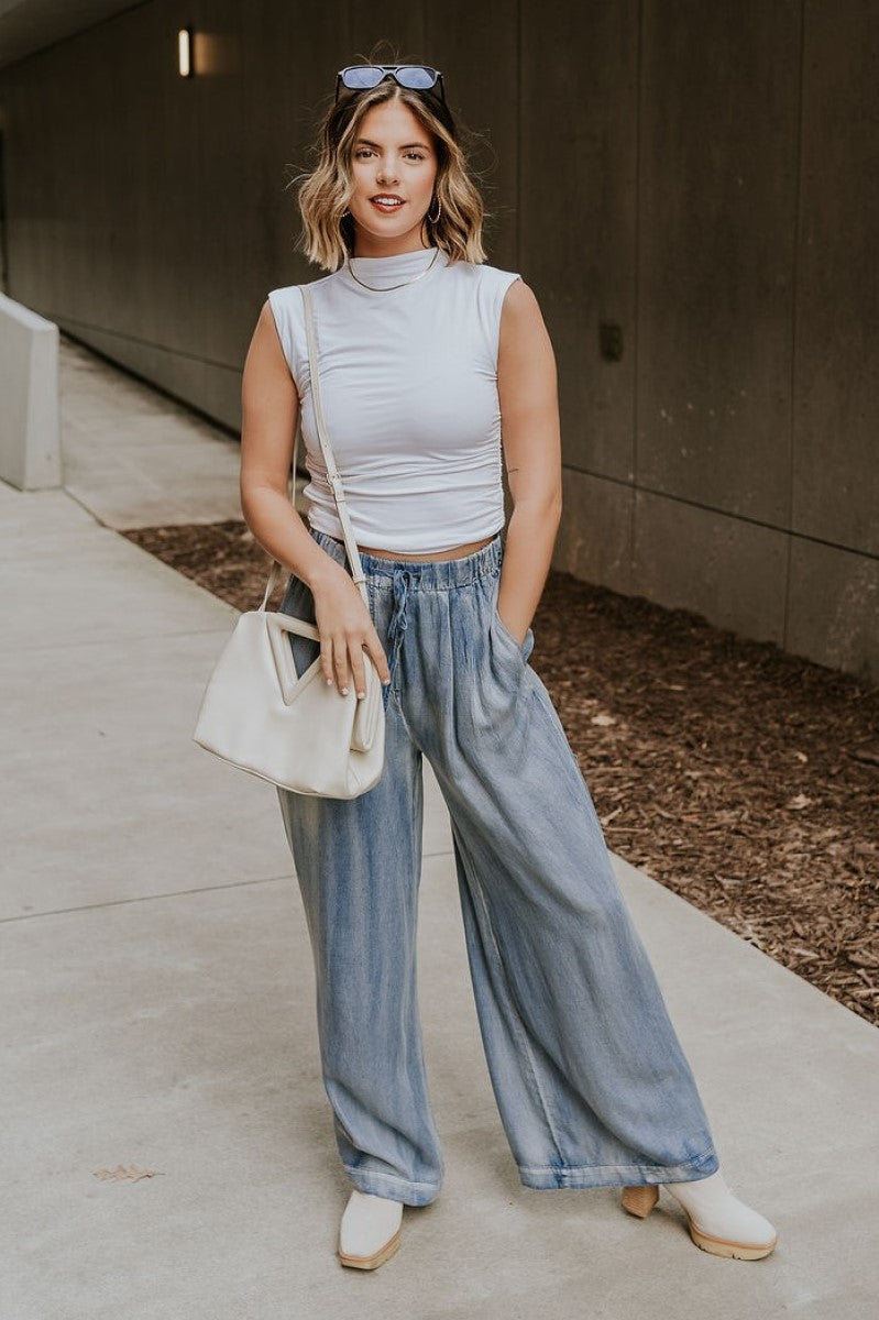Full body front view of model wearing the Kelly Blue Wide Leg Drawstring Pants that have blue denim-washed light weight fabric, two front slit pockets, an elastic waistband with a drawstring tie, and wide pant legs.