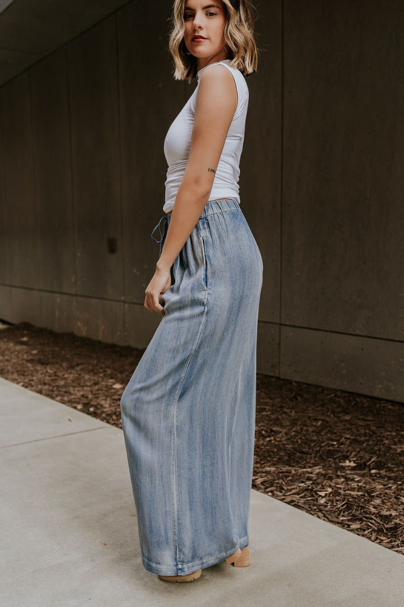 Full body side view of model wearing the Kelly Blue Wide Leg Drawstring Pants that have blue denim-washed light weight fabric, two front slit pockets, an elastic waistband with a drawstring tie, and wide pant legs.