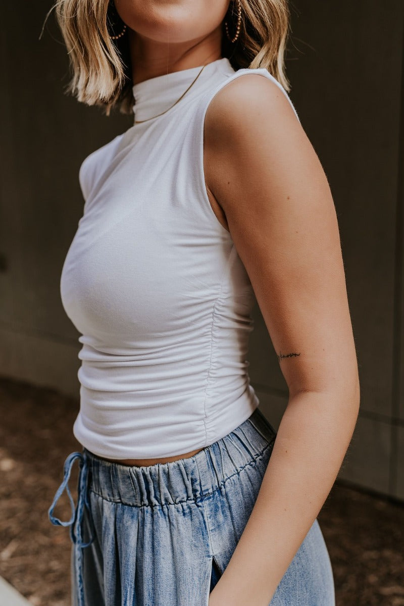 Side view of model wearing the Rebekah White Ruched Mock Neck Tank Top that has double layer white knit fabric, ruched side details, a high neckline, and a sleeveless design.